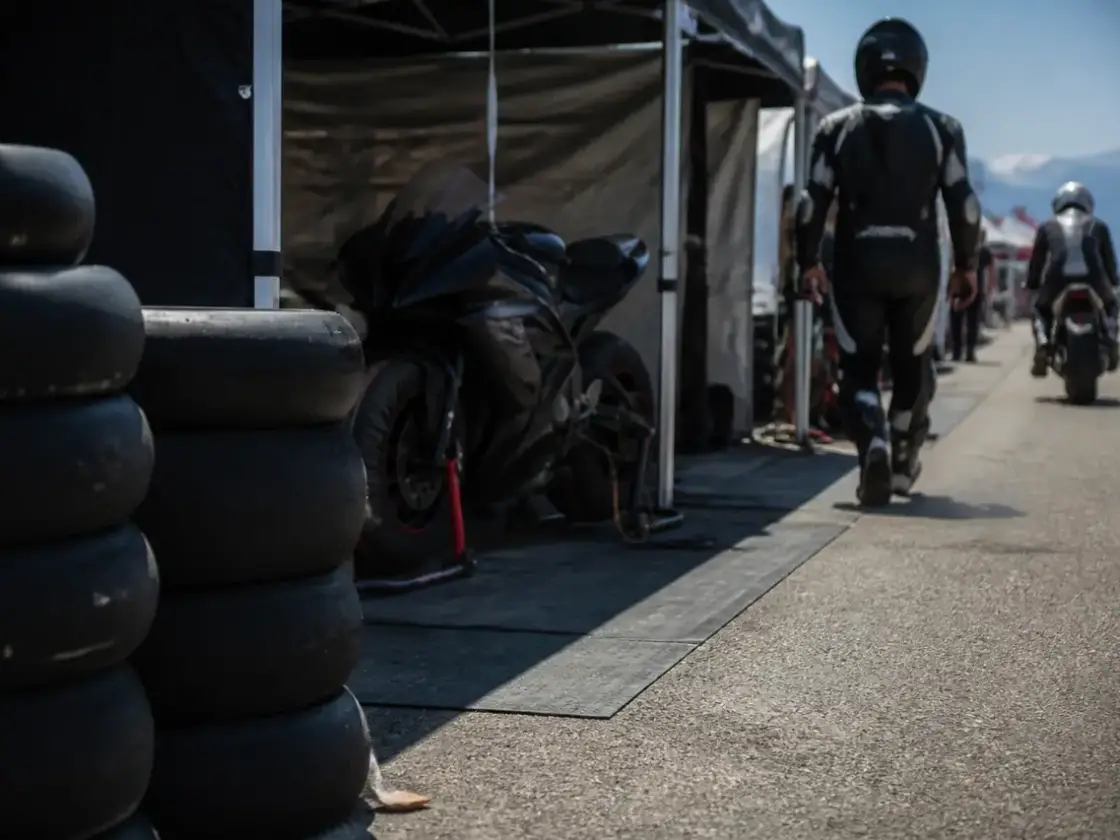 Voie des stands d’un circuit un jour de course, pneus empilés devant une tente ouverte avec une moto sur béquille, personnes de dos et floues