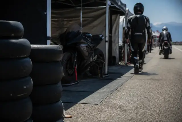 Voie des stands d’un circuit un jour de course, pneus empilés devant une tente ouverte avec une moto sur béquille, personnes de dos et floues