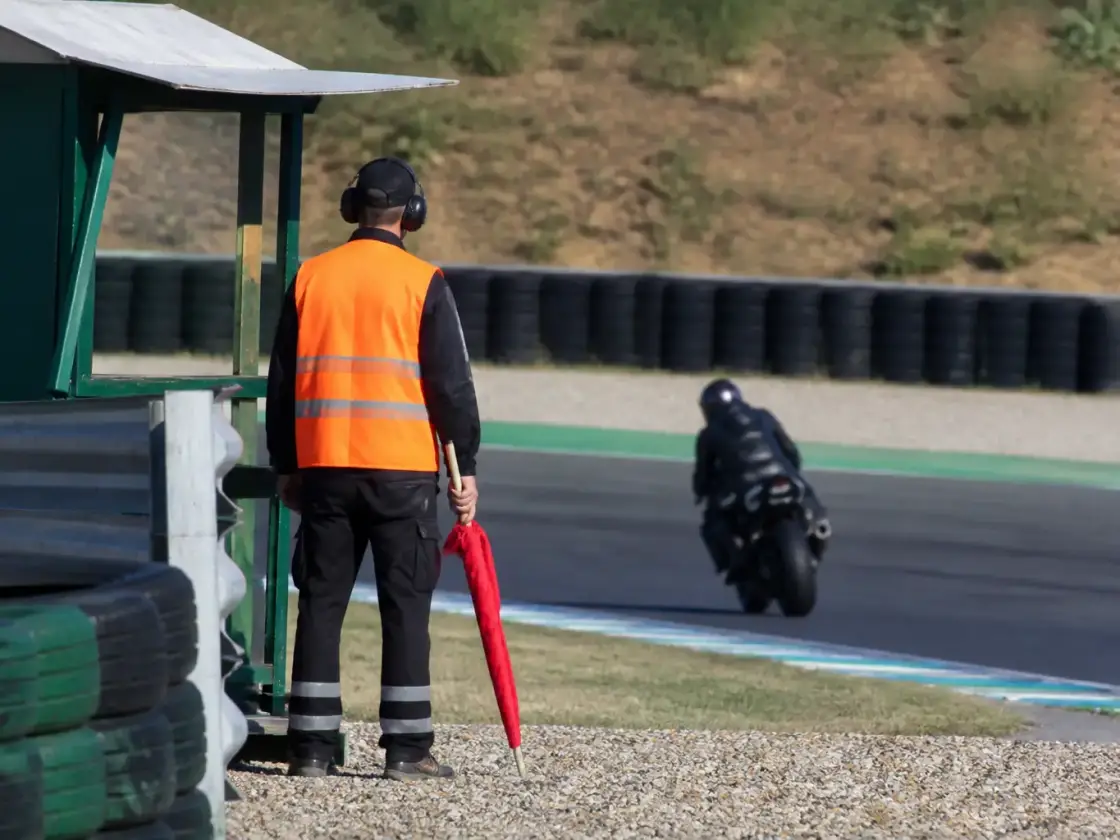 Commissaire de piste en gilet de signalisation debout près d’un poste de bord de piste d’un circuit, drapeau roulé à la main, aucun visage ni inscription lisible
