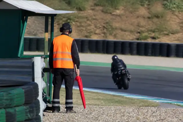 Commissaire de piste en gilet de signalisation debout près d’un poste de bord de piste d’un circuit, drapeau roulé à la main, aucun visage ni inscription lisible