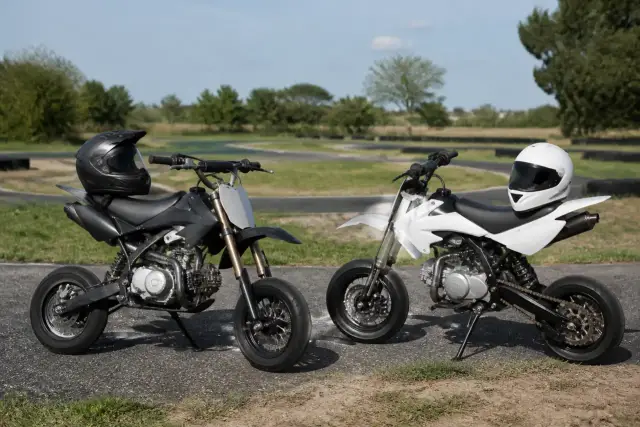 Deux petites motos de type pit bike garées sur un petit circuit de kart, casques posés sur les selles, herbe et bitume