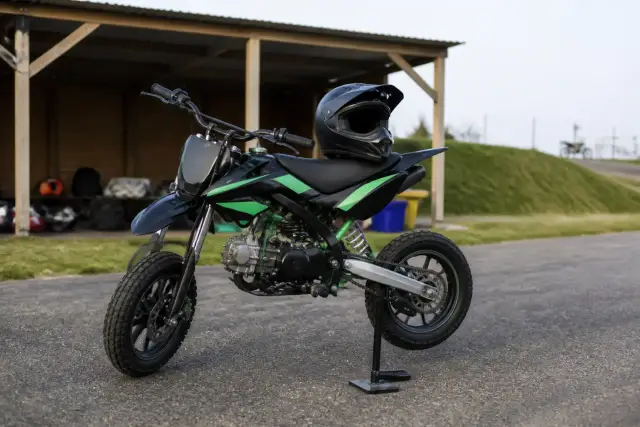 Petite moto de type pit bike garée dans un paddock de piste de loisir, casque posé sur la selle, aucun lieu ni inscription reconnaissable