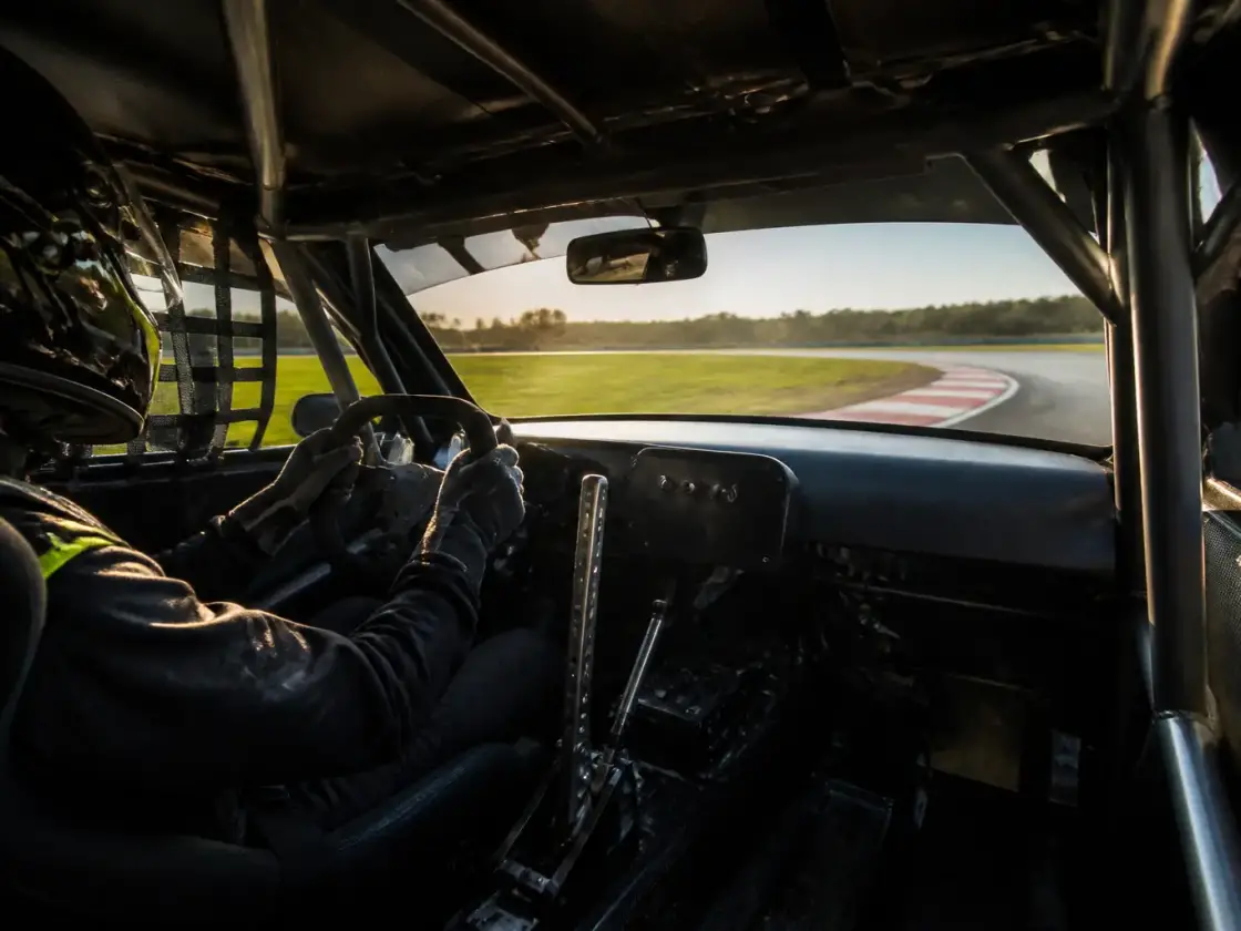 Pilote casqué au volant d’une voiture de piste au dessin inventé, habitacle dépouillé avec arceau de sécurité et levier de vitesses, virage et vibreur au loin