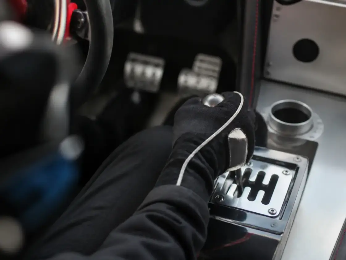Main gantée sur un levier de vitesses à grille en H dans une voiture de piste au dessin inventé, pieds sur le pédalier en arrière-plan flou