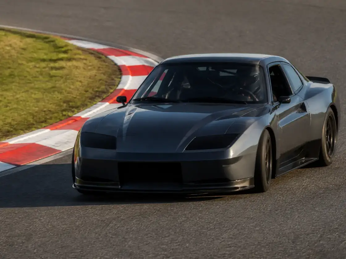Voiture de piste gris anthracite qui aborde un virage serré sur un circuit, vue de trois quarts avant, vibreur rayé rouge et blanc à la corde