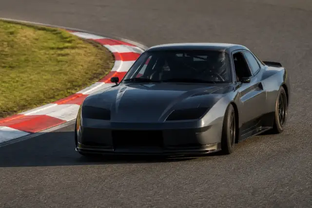 Voiture de piste gris anthracite qui aborde un virage serré sur un circuit, vue de trois quarts avant, vibreur rayé rouge et blanc à la corde