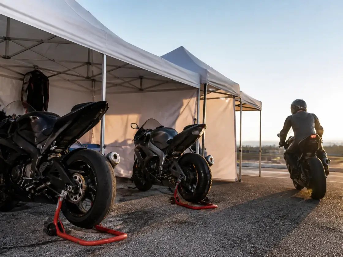 Vue large d’un paddock avec des motos sur béquille d’atelier, des tentes blanches et un ciel clair de début de journée