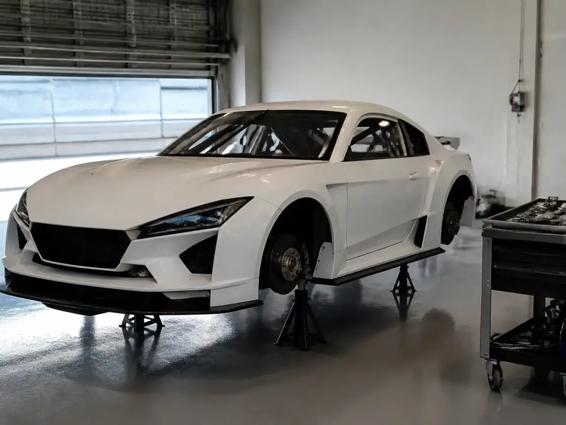 Voiture de piste blanche posée sur des chandelles dans un box, roues avant déposées, disque de frein visible, chariot d’outils à côté