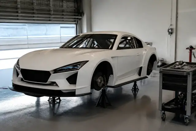 Voiture de piste blanche posée sur des chandelles dans un box, roues avant déposées, disque de frein visible, chariot d’outils à côté
