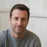 Portrait de Sacha Tissandier, cheveux châtains courts, barbe de trois jours, pull gris, sourire discret, mur clair flou en arrière-plan