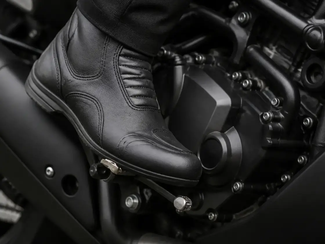 Pied gainé de cuir posé sur le sélecteur d’une moto, cadre et moteur noirs flous en arrière-plan