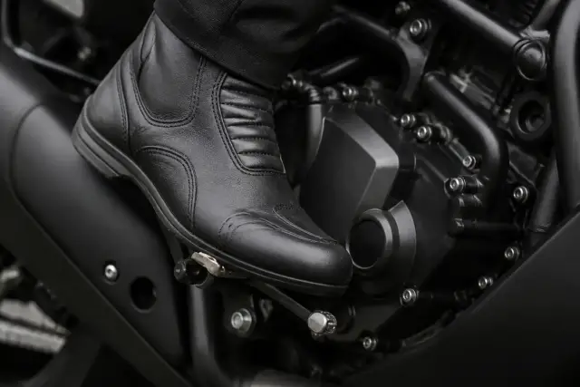 Pied gainé de cuir posé sur le sélecteur d’une moto, cadre et moteur noirs flous en arrière-plan