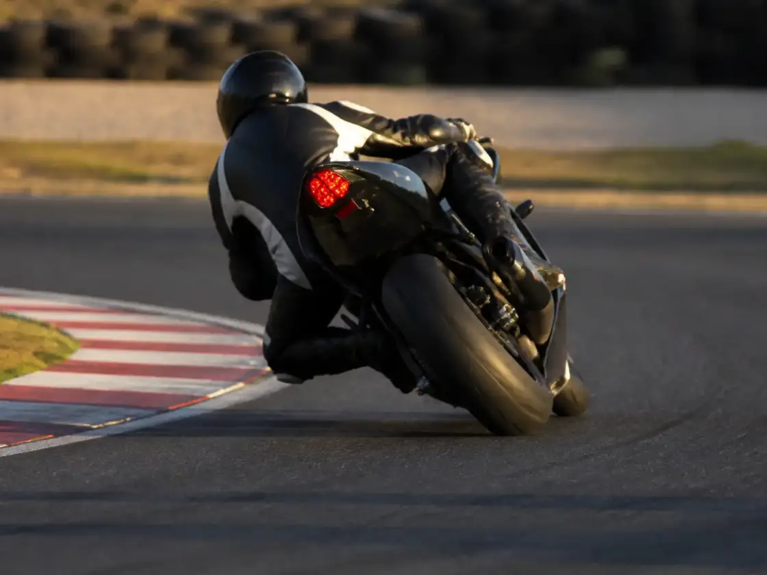 Moto vue de dos à la sortie d’un virage serré sur un circuit, pilote casqué penché, bitume sec et vibreur rayé rouge et blanc