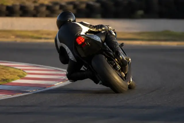Moto vue de dos à la sortie d’un virage serré sur un circuit, pilote casqué penché, bitume sec et vibreur rayé rouge et blanc