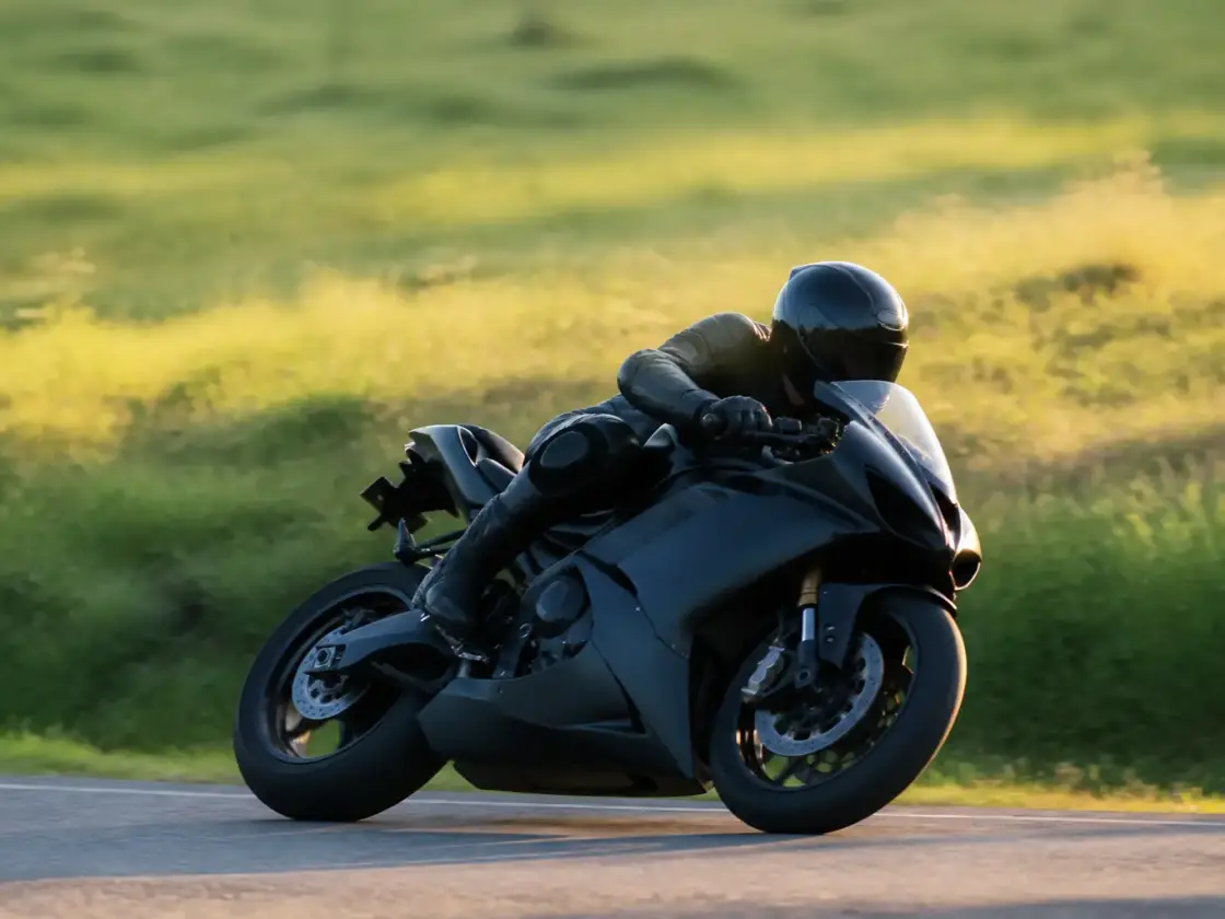 Moto penchée en courbe vue de côté sur une petite route de campagne, pilote casqué non identifiable, lumière rasante