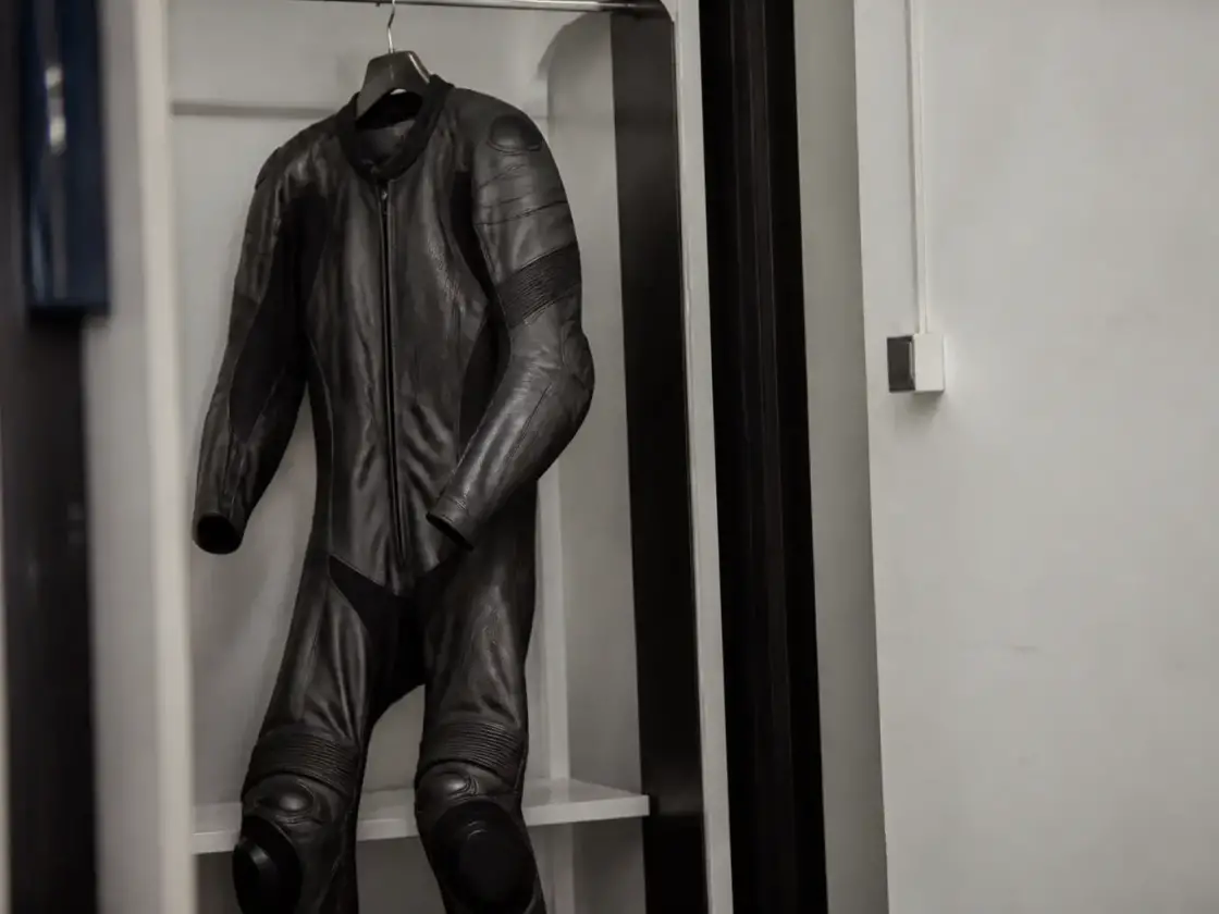 Combinaison de cuir de moto sombre, pendue à un cintre dans un vestiaire neutre, coudes et genoux renforcés visibles