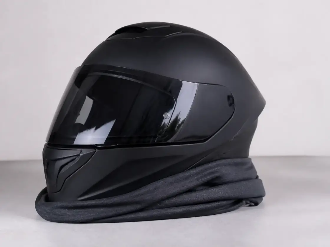 Tour de cou en tissu technique gris posé sur un casque de moto intégral, fond uni clair