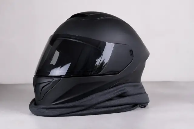Tour de cou en tissu technique gris posé sur un casque de moto intégral, fond uni clair